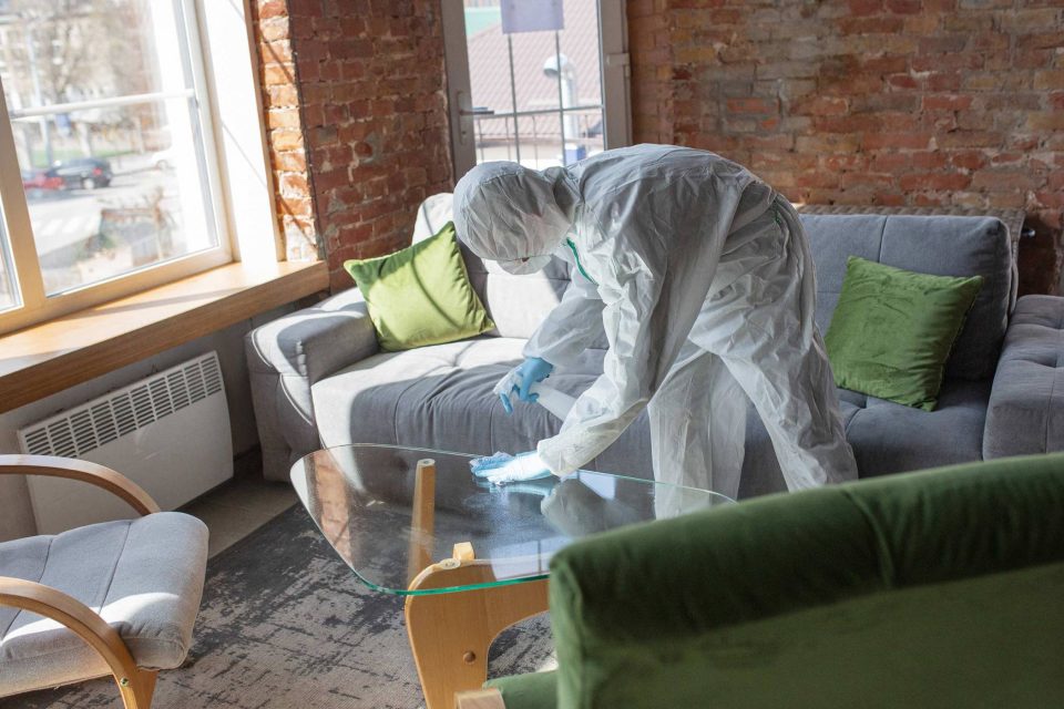 Person in protective suit doing biohazard cleanup on living rooms glass table.